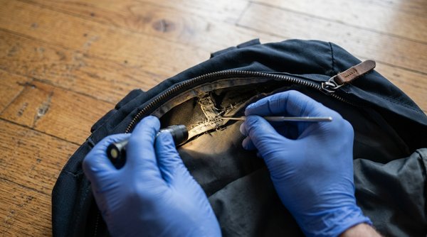 Gloved technician inspecting a backpack zipper seam during a bed bug inspection, showing a common but overlooked hiding spot.